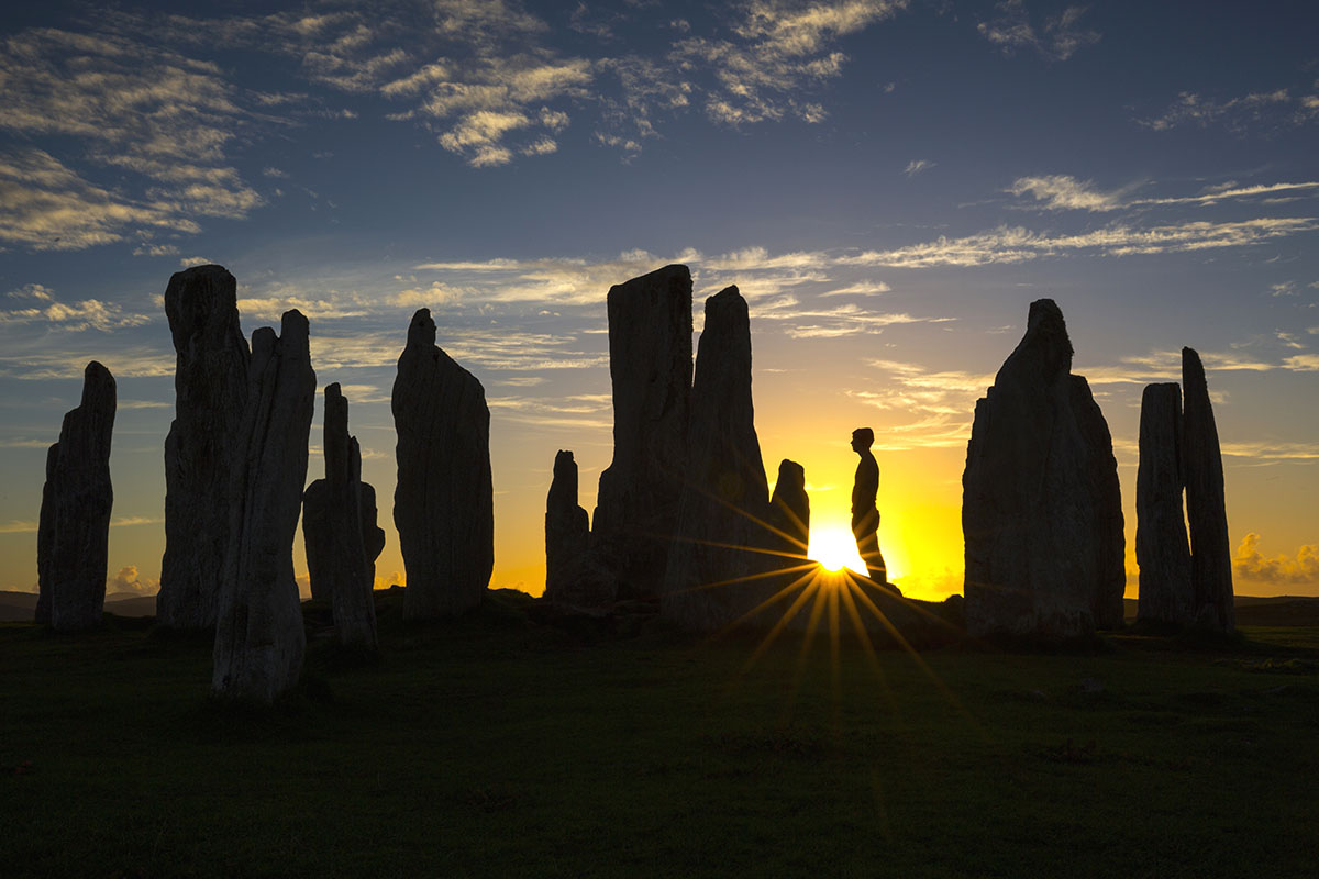 A person stands among standing stones with the sun rising behind