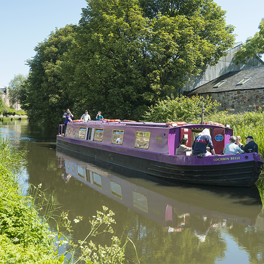 7 people on purple canal boat, sailing along water with green trees