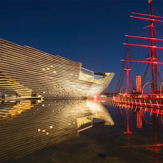 V&A Dundee lit up at night, next to RRS Discovery lit up red.