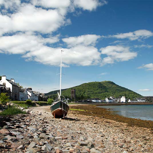 a boat sits on a stony beach, with a row of houses in the background